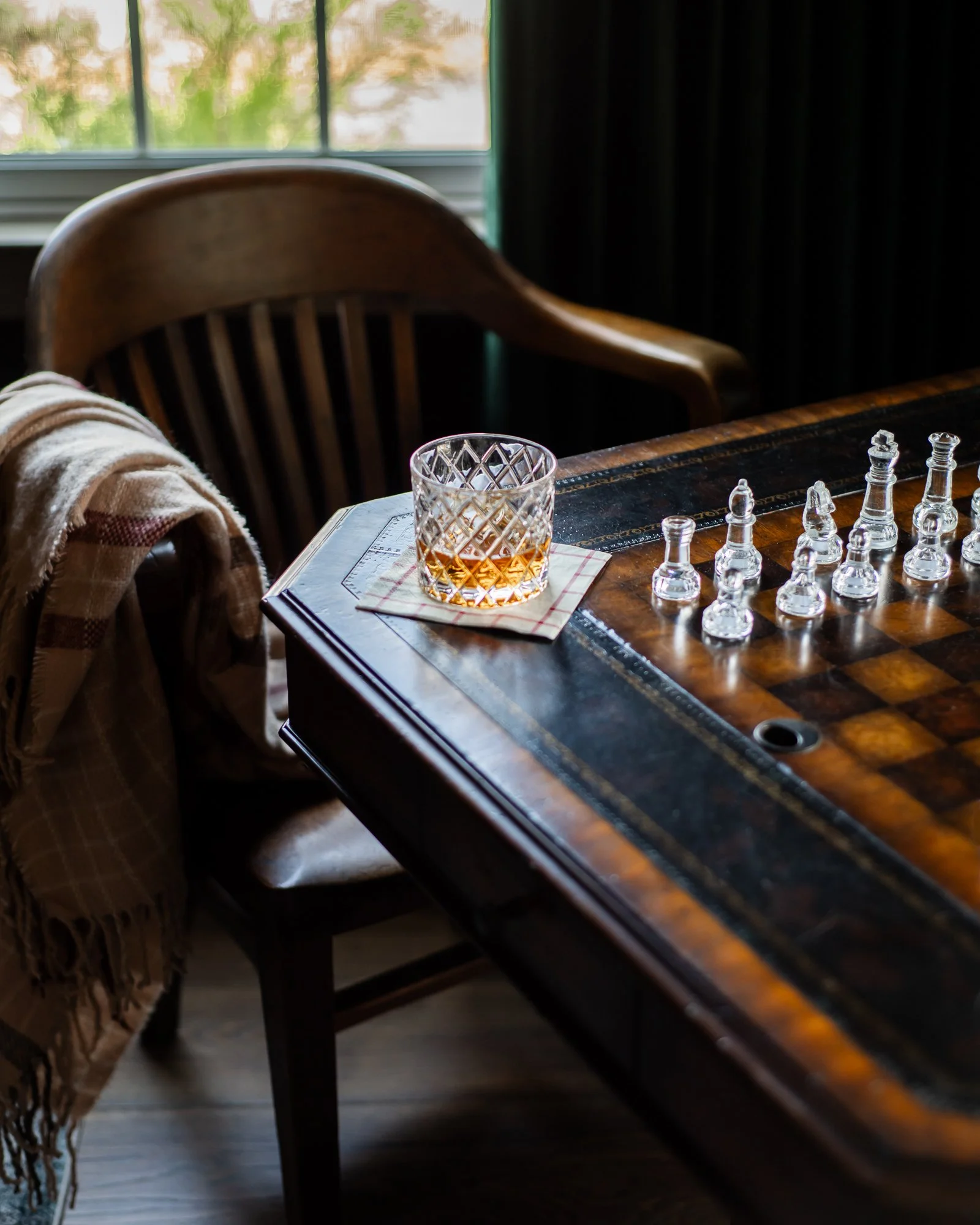 A chessboard on a wooden table with a glass of whiskey on a checkered napkin, near a wooden chair with a plaid blanket, inside a room with a window showing green trees outside.