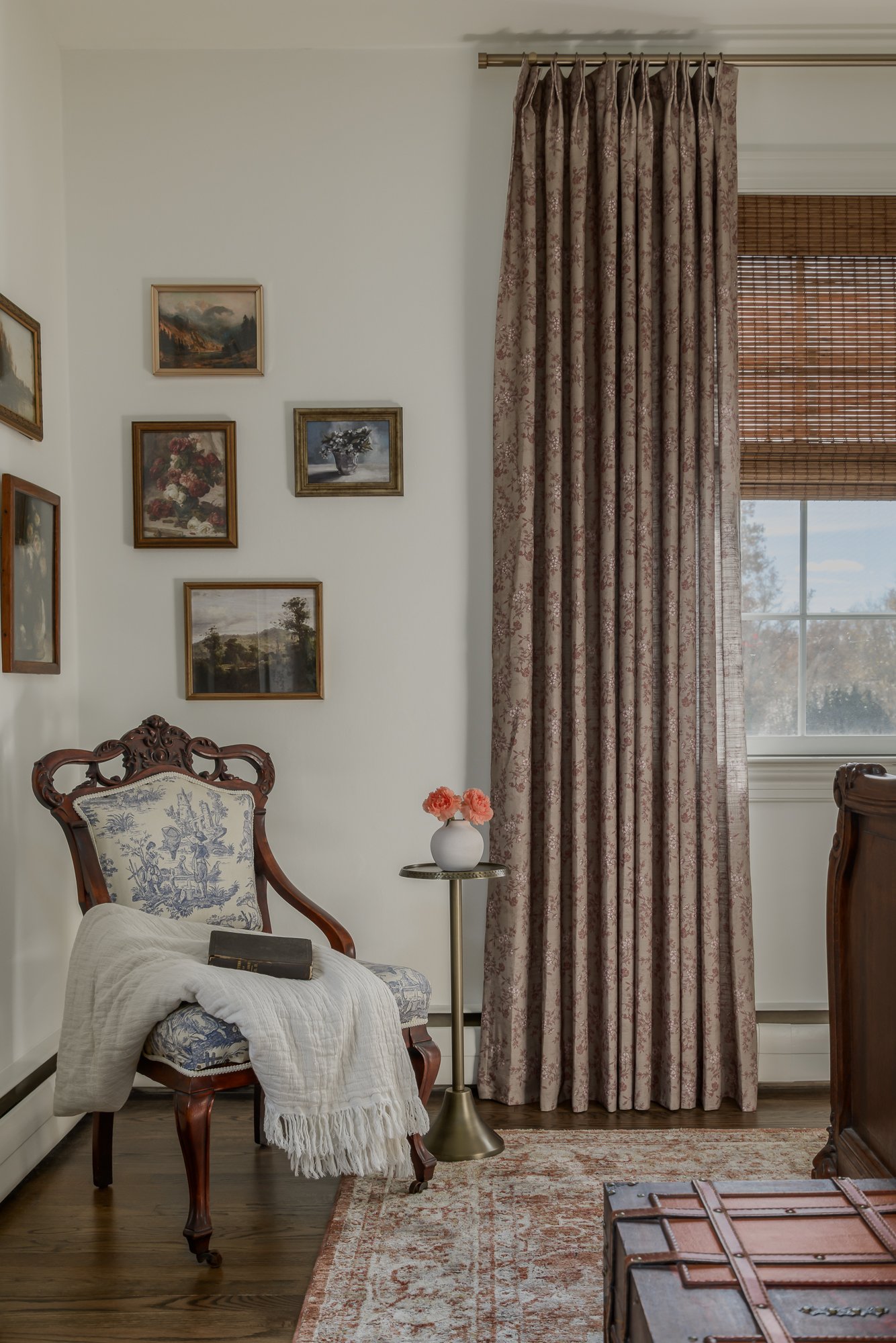 A corner of a vintage-style room with a wooden chair with upholstered floral fabric, a white throw blanket, and a closed book on the chair's seat. Next to the chair is a small round metal side table with pink flowers in a white vase. Behind is a wind