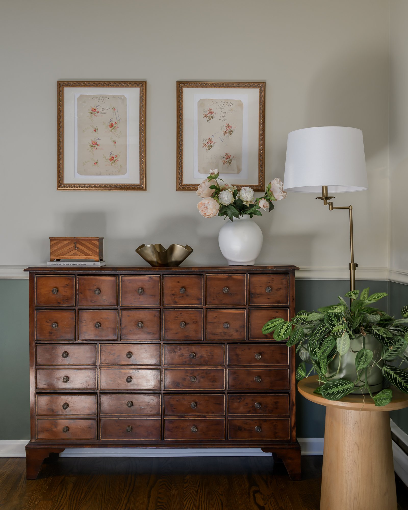 A vintage wooden cabinet with multiple small drawers, decorated with a white vase of pink and white flowers, a brass bowl, and a wooden box on top, against a wall with two framed botanical prints, a tall white floor lamp with a white shade, and a pot
