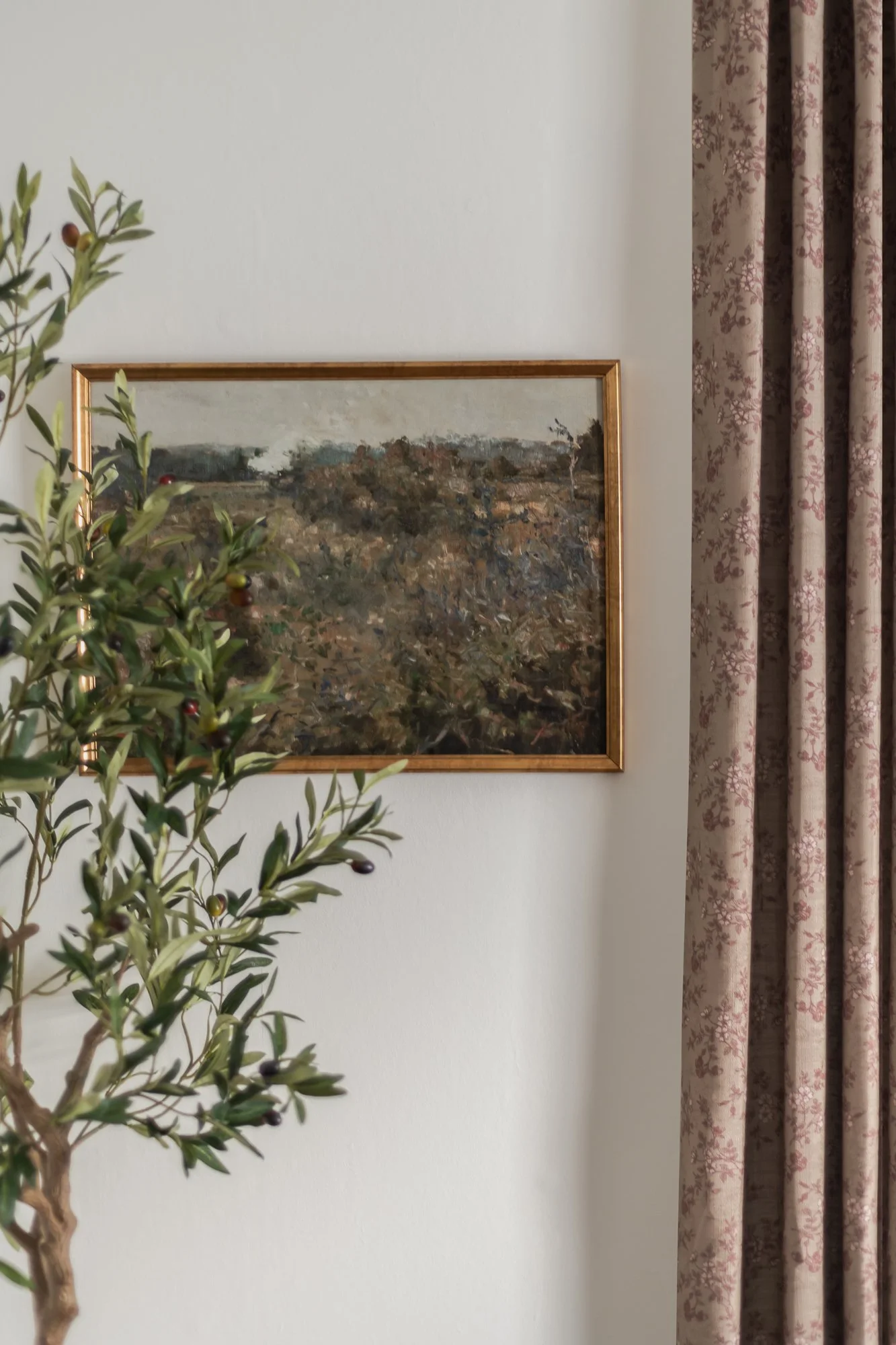 Close-up of a decorative wall with an abstract landscape painting in a gold frame, a leafy plant with small dark berries on the left, and patterned curtains on the right.
