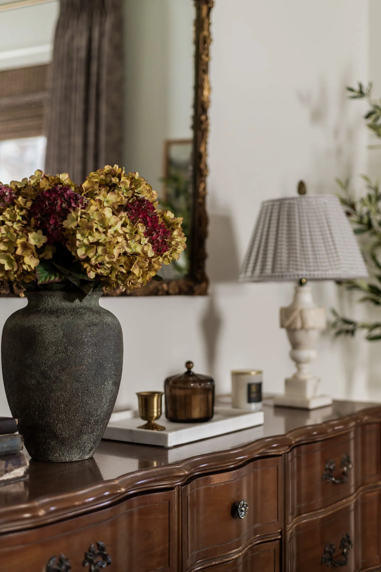A wooden sideboard with a textured ceramic vase filled with dried hydrangea flowers, a small gold cup, a dark glass jar, candles, a table lamp, and a mirror in a well-lit living room.