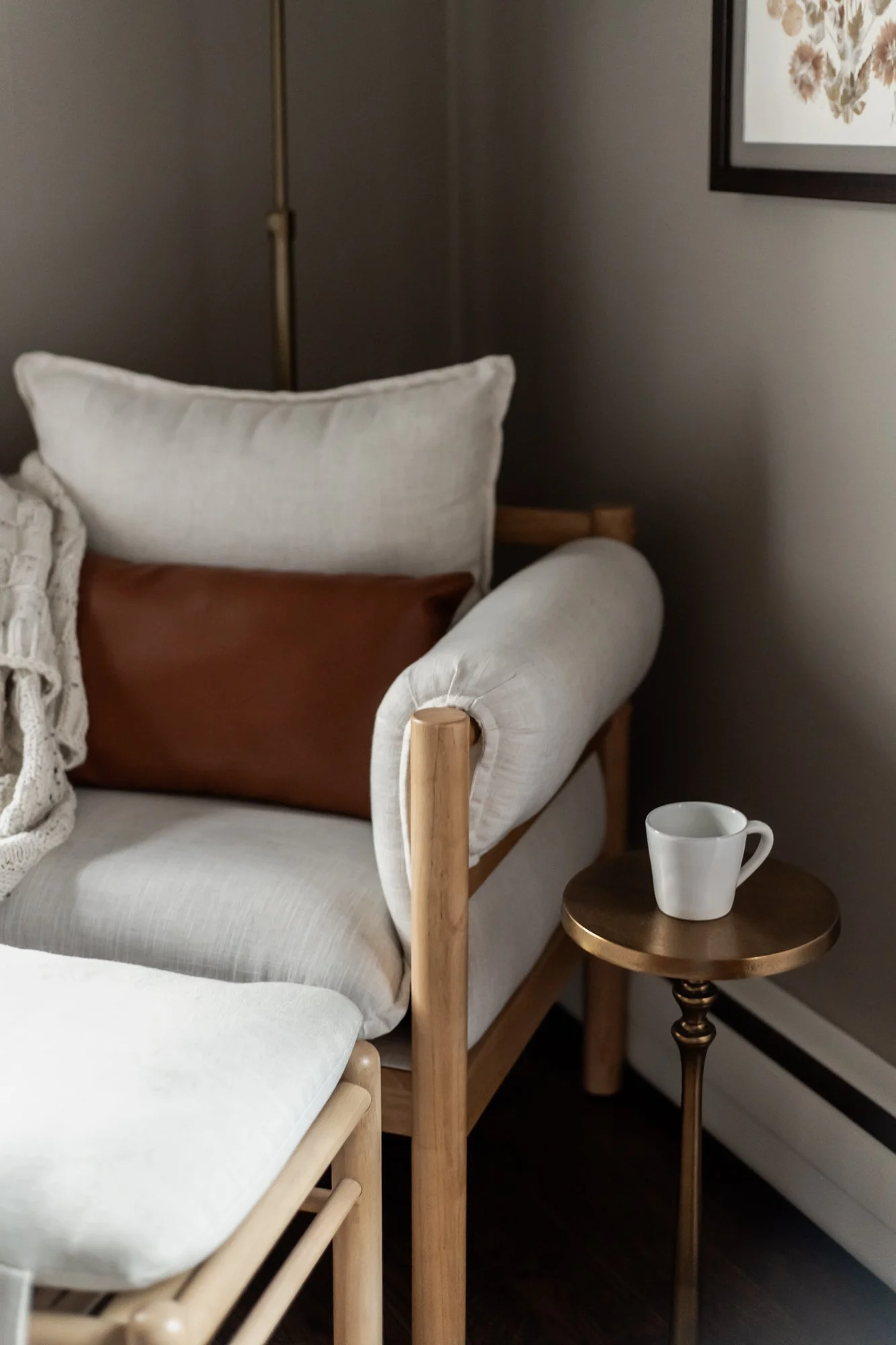 A cozy living room corner with a cream-colored armchair, a side table holding a white mug, and a framed floral artwork on the wall.