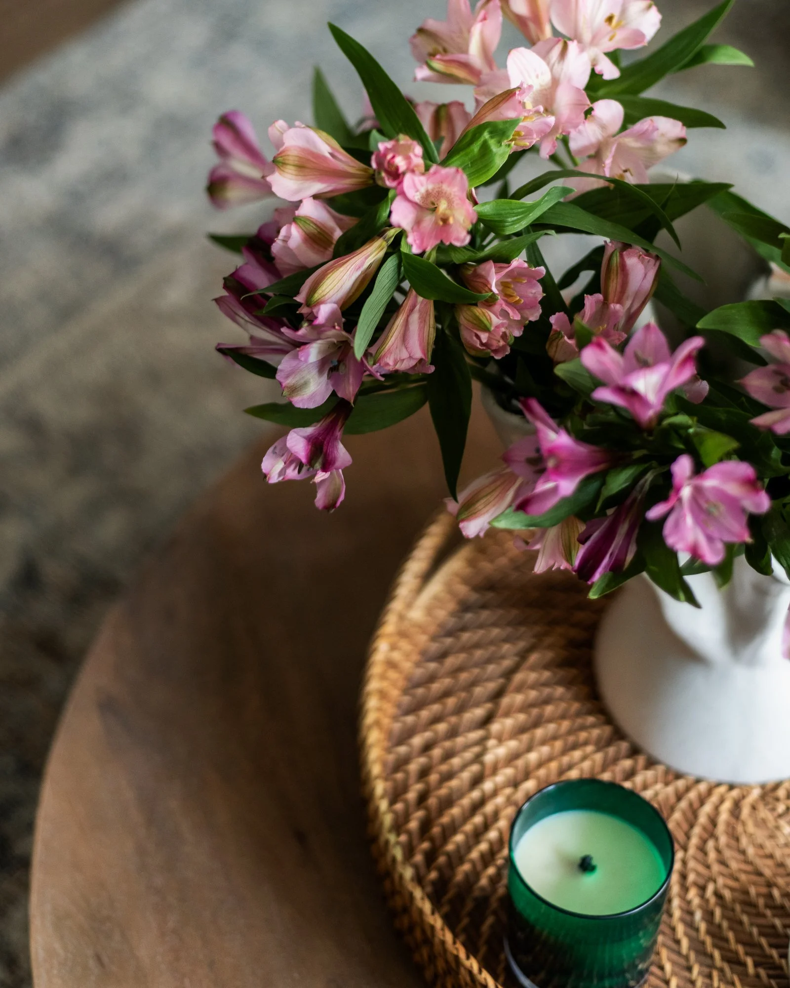 A white vase with pink flowers and green leaves on a woven tray, with a green candle nearby, on a wooden surface.