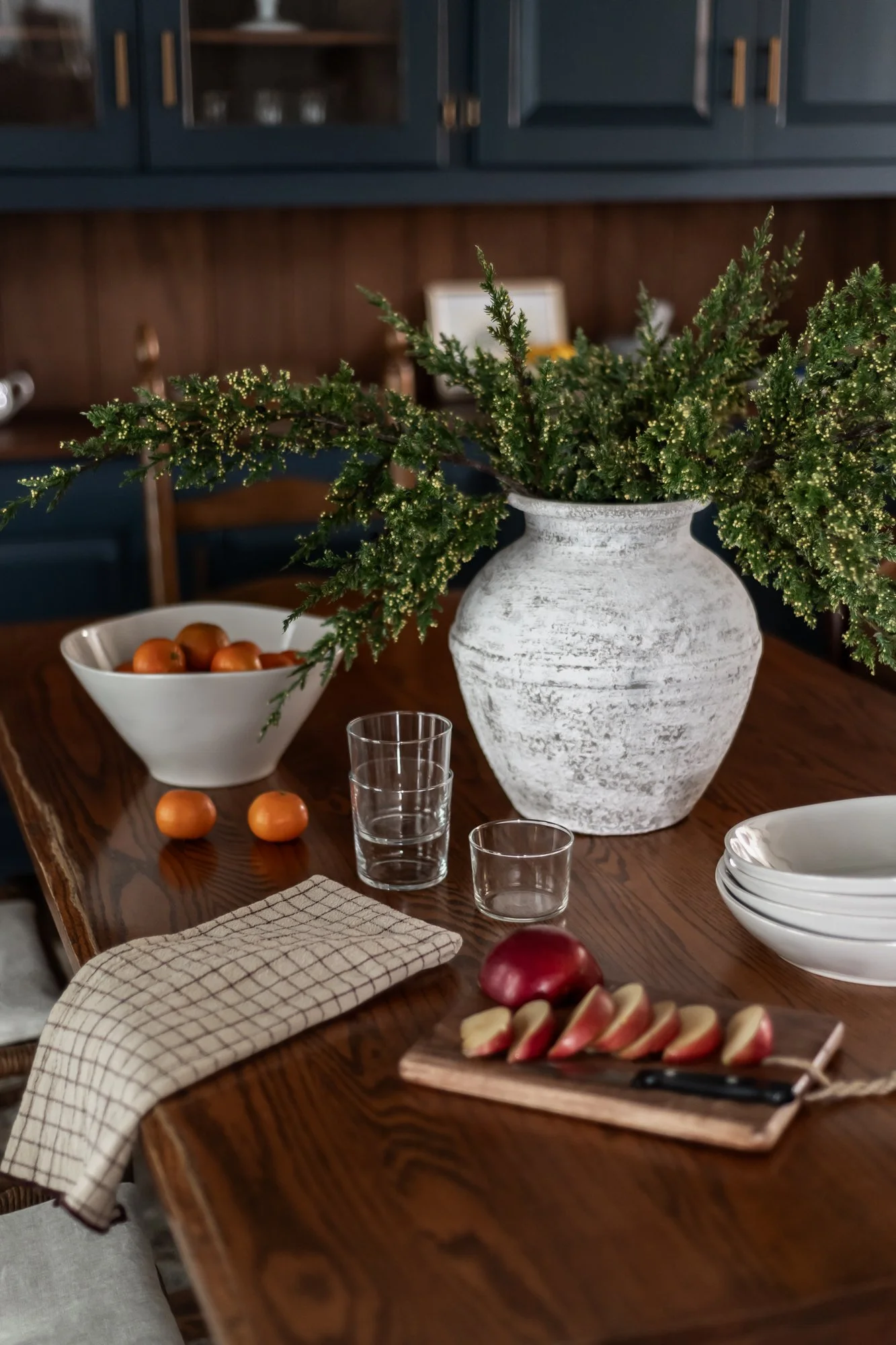 A wooden dining table set with a large white vase filled with greenery, a bowl of small fruit, a sliced apple on a wooden cutting board, stacked white bowls, drinking glasses, and a checkered napkin.