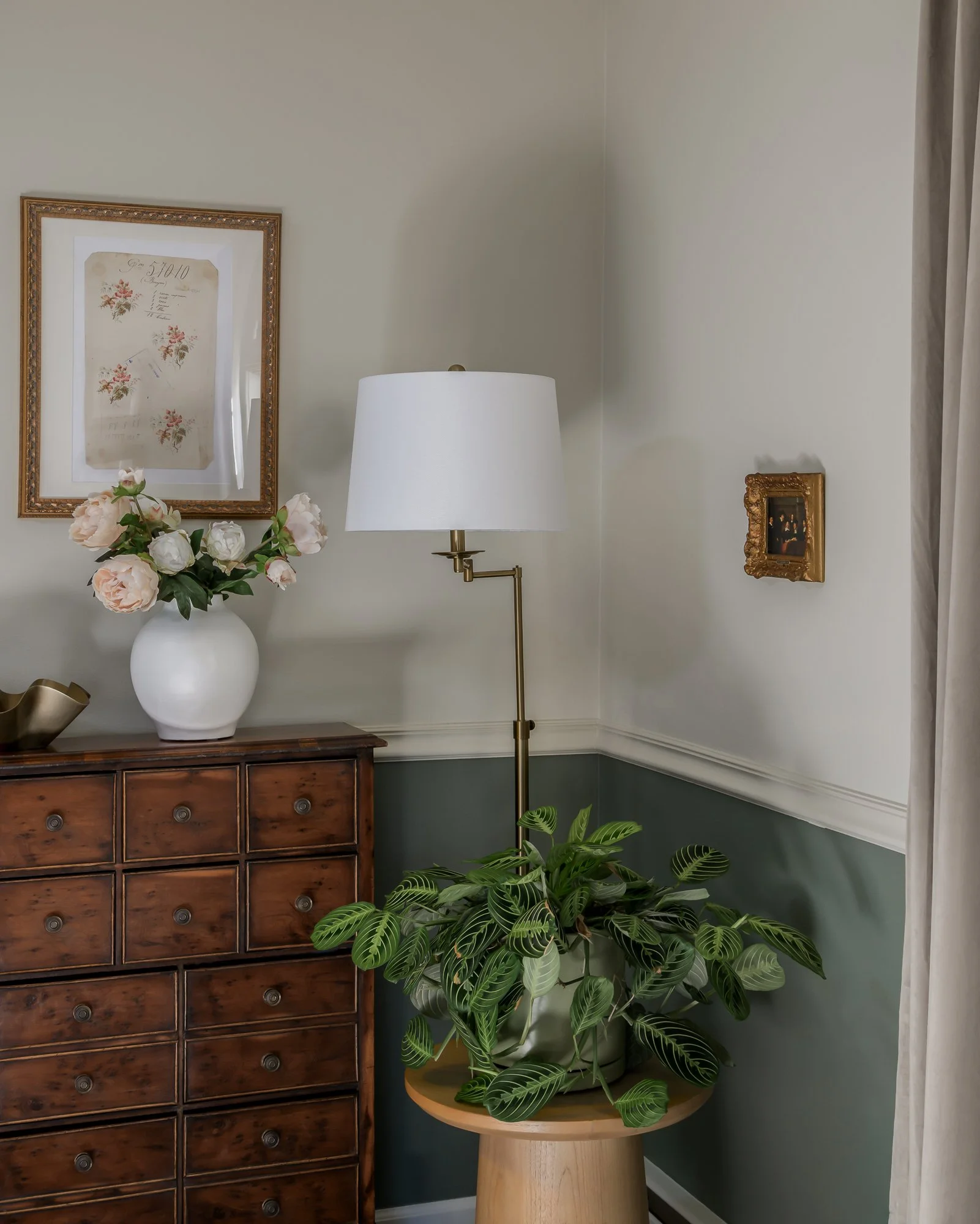 Interior corner with a wooden dresser, a white vase with pink and white peonies, a gold framed wall art, a brass floor lamp with a white shade, and a potted plant on a wooden stand next to cream-colored curtains.