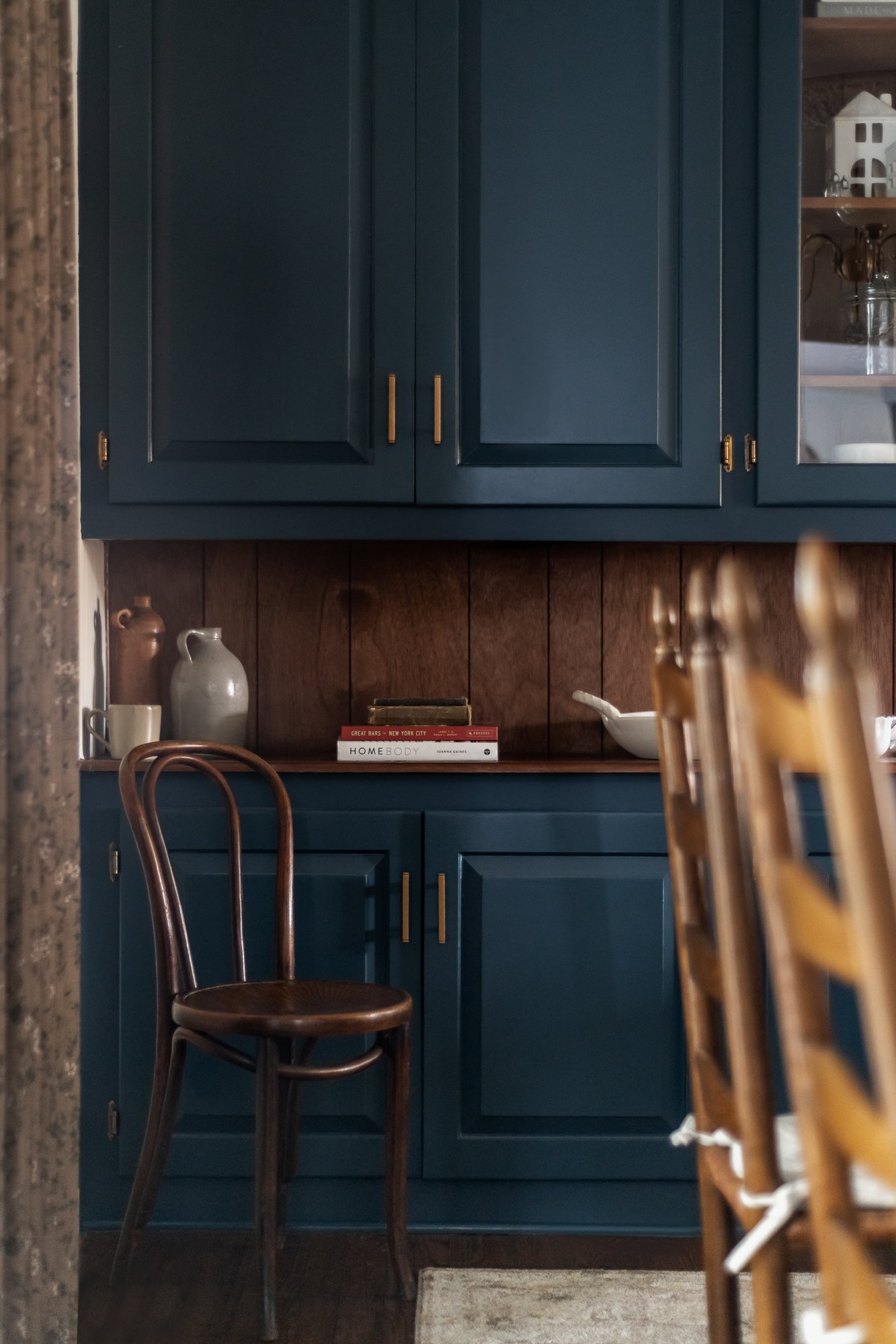 A blue kitchen cabinet with gold handles, a wooden countertop, and a dark wooden chair with a curved back in front of the cabinet. On the countertop are two vases, a stack of books, and a white bowl. To the right, there are wooden chairs with vertica
