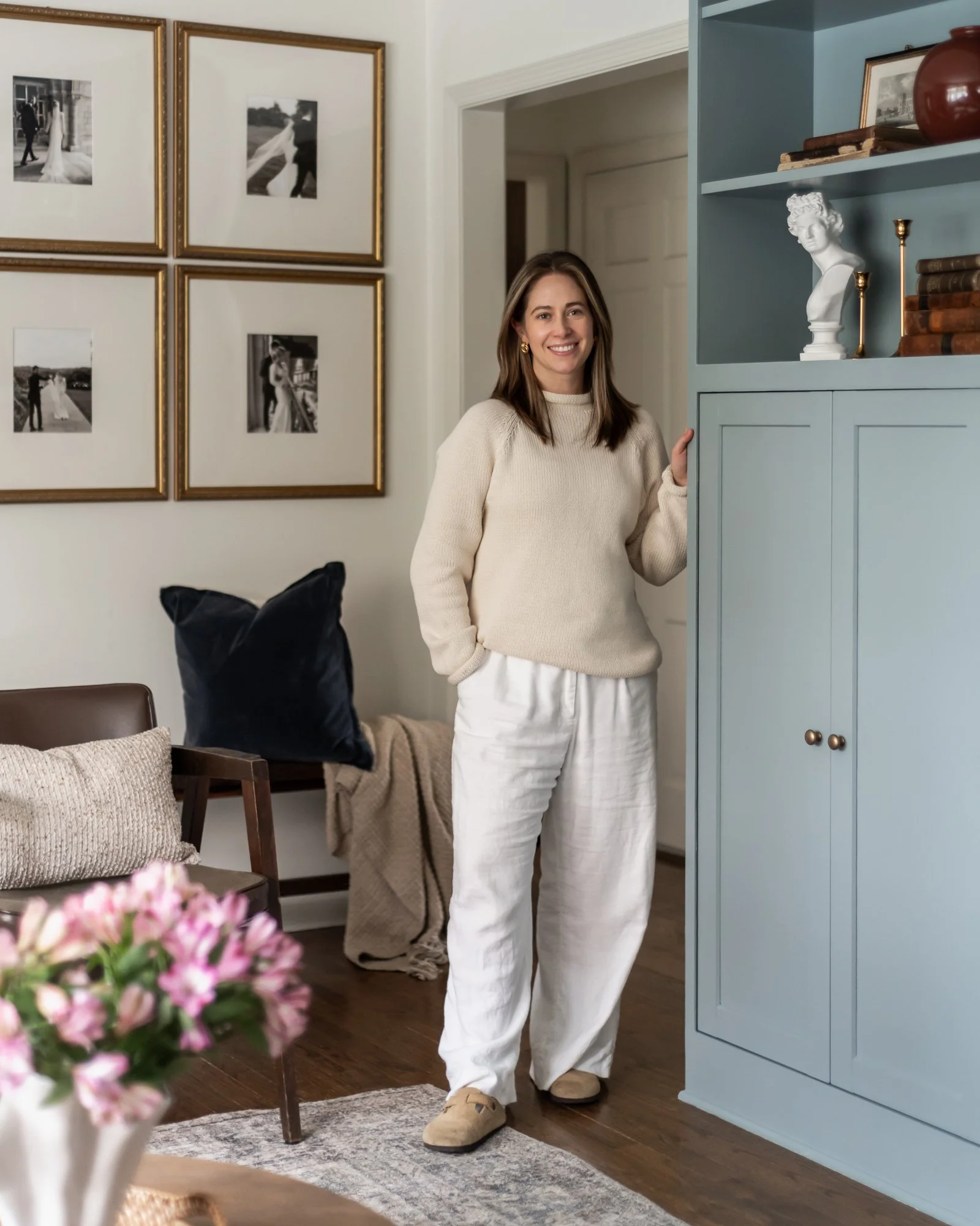 A woman wearing a cream sweater and white pants, standing in a living room, smiling at the camera, near a blue cabinet with decorative items, against a wall with black-and-white photographs in gold frames.