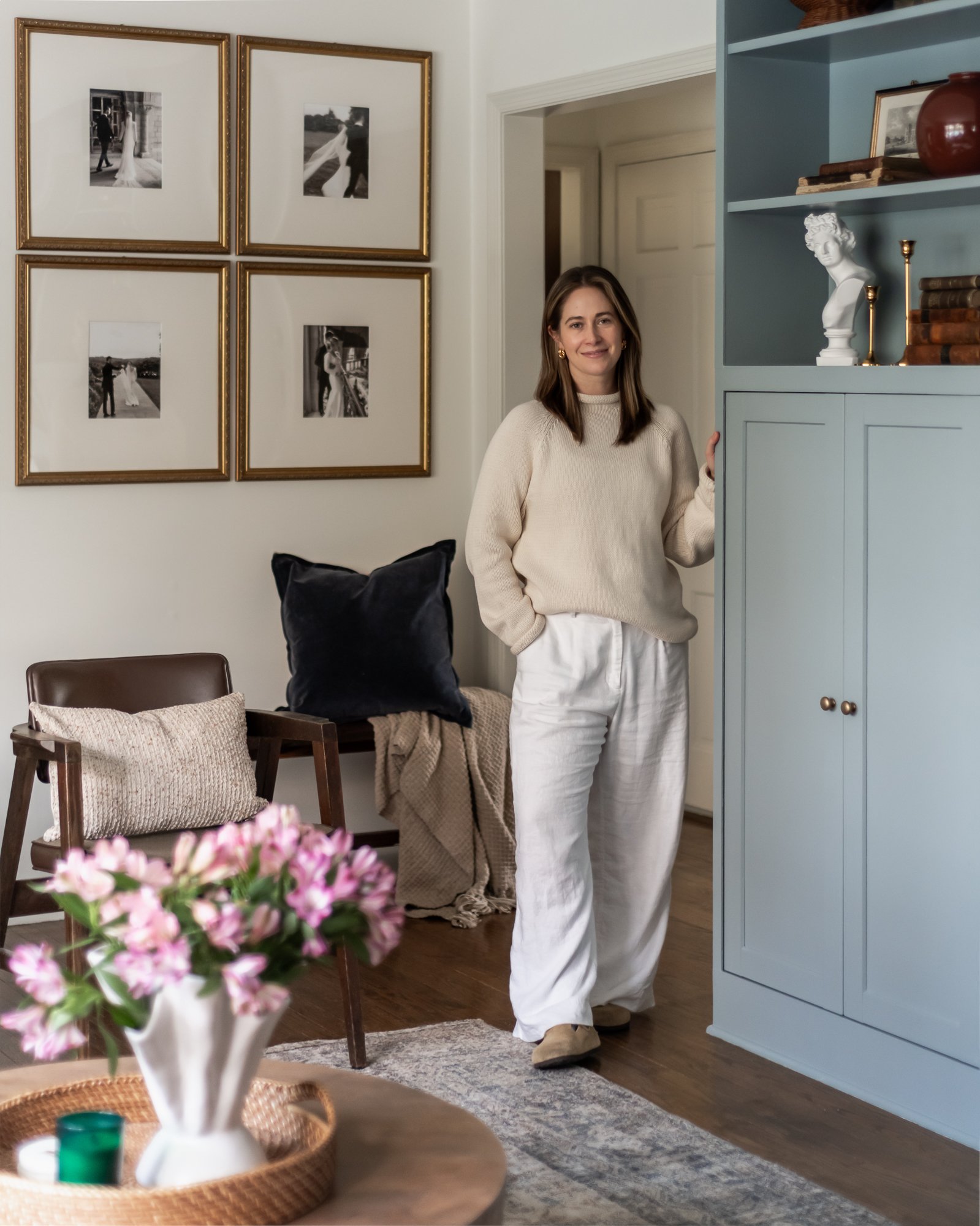 A woman standing inside a living room, wearing a beige sweater and white pants, near a light blue cabinet with decorative items on top, including a white bust sculpture and a red vase.