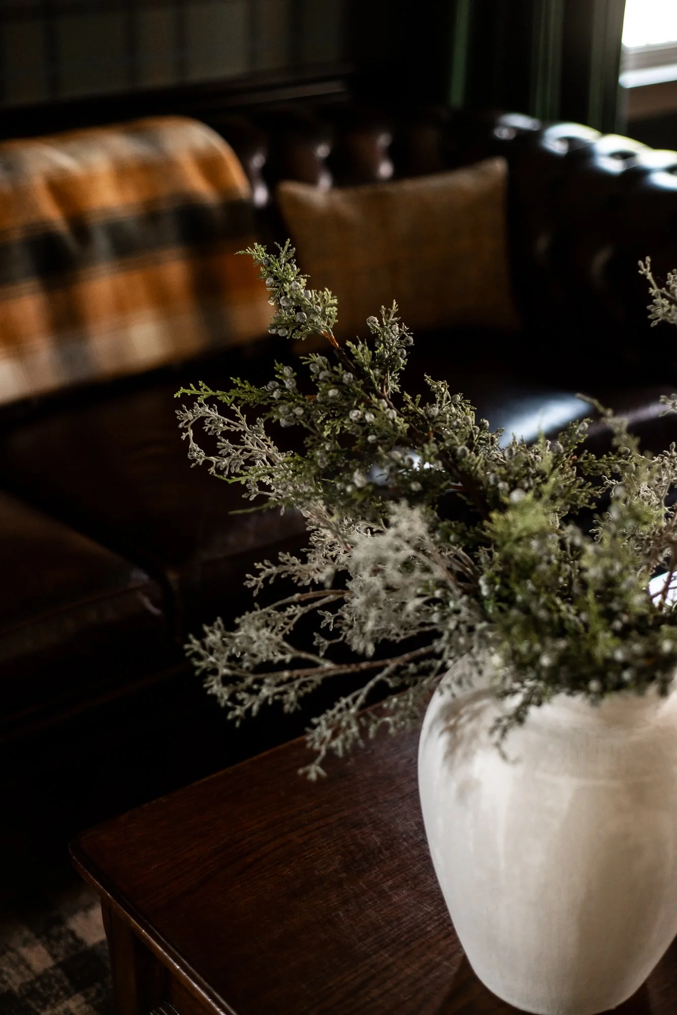 A white vase with green and white faux greenery on a wooden table in a dimly lit living room with leather chairs