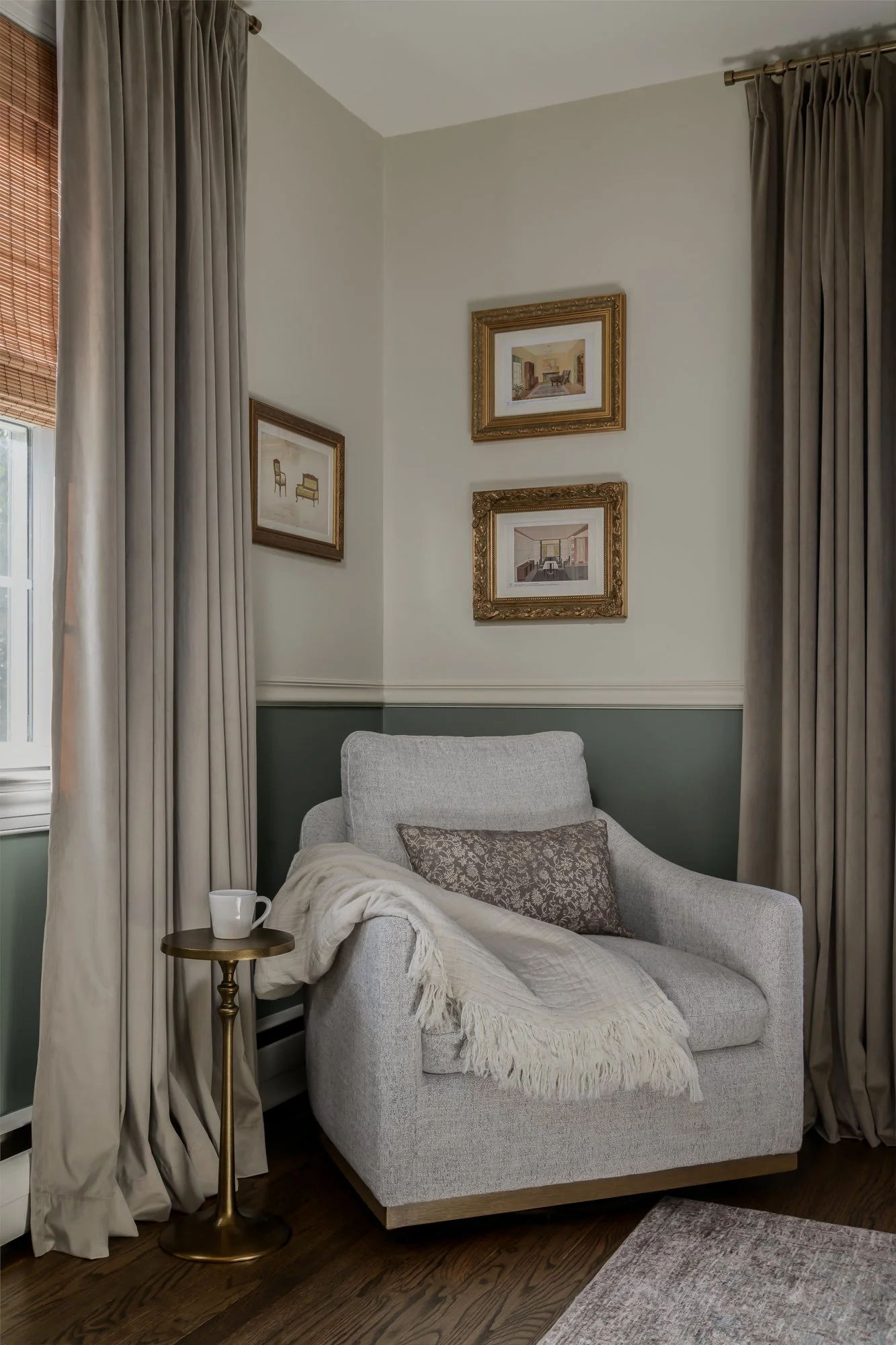 A cozy corner with a light grey armchair draped with a cream throw blanket and a decorative pillow, positioned next to a small round gold side table holding a white mug. The corner features cream and taupe curtains, framed artwork on the wall, a wind