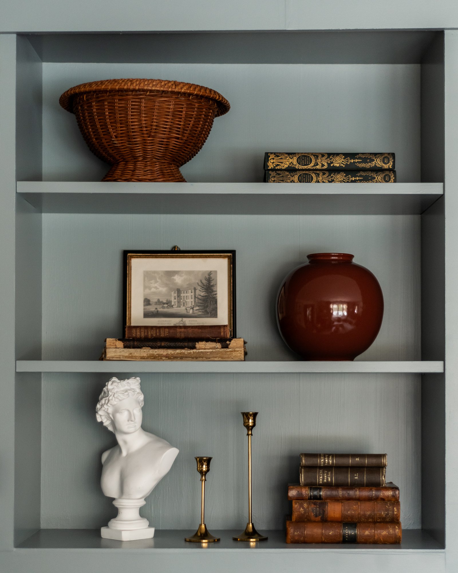 Decorative bookshelf with woven basket, vintage books, framed picture, large ceramic pot, classical bust, brass candlesticks, and more antique books.