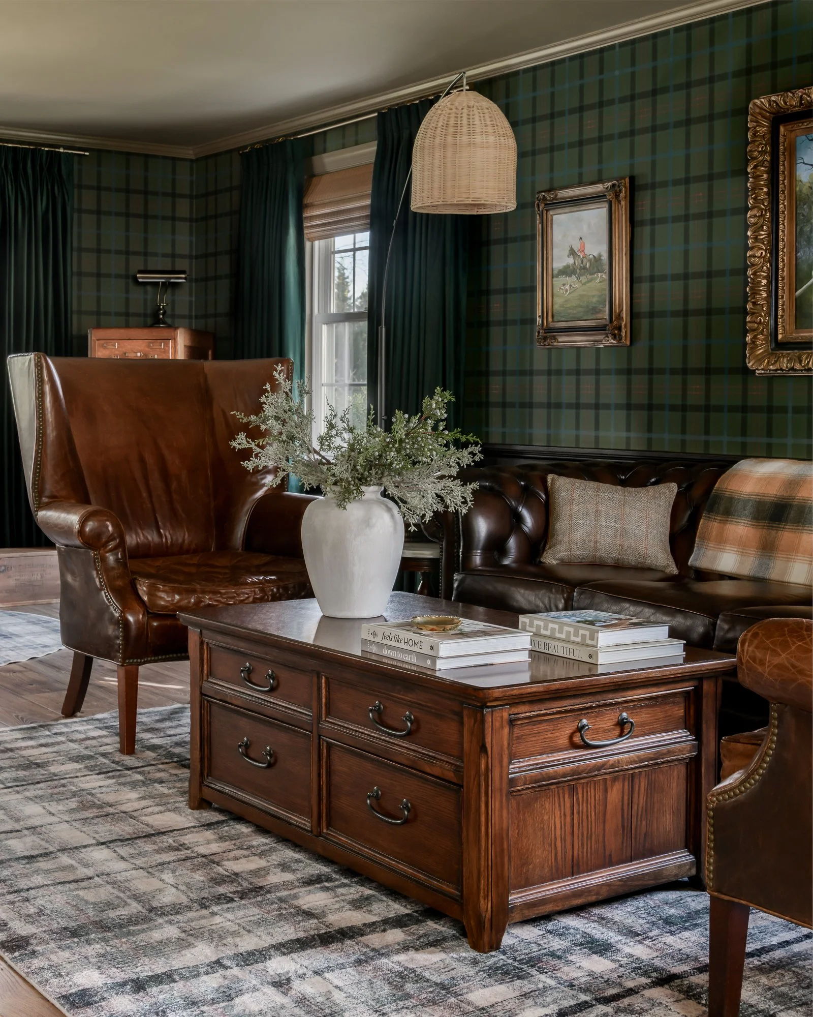 Living room with green plaid wallpaper, dark green curtains, wooden coffee table with a white vase and green foliage, leather armchair and sofa, framed artwork, and a patterned rug.