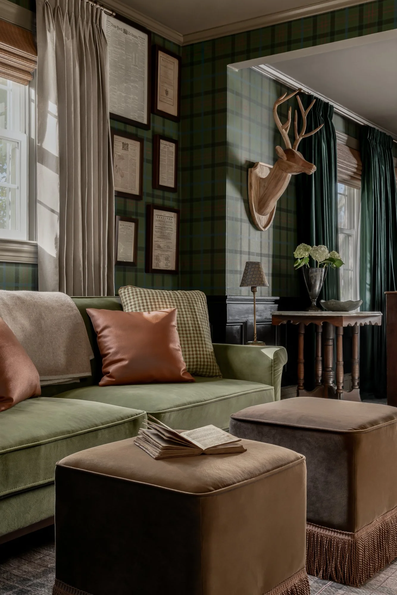A cozy living room with a green upholstered sofa adorned with brown and patterned cushions, two beige ottomans, a side table with a lamp and a vase of white flowers, a window with curtains, and a wall decorated with framed newspaper articles and a deer head.