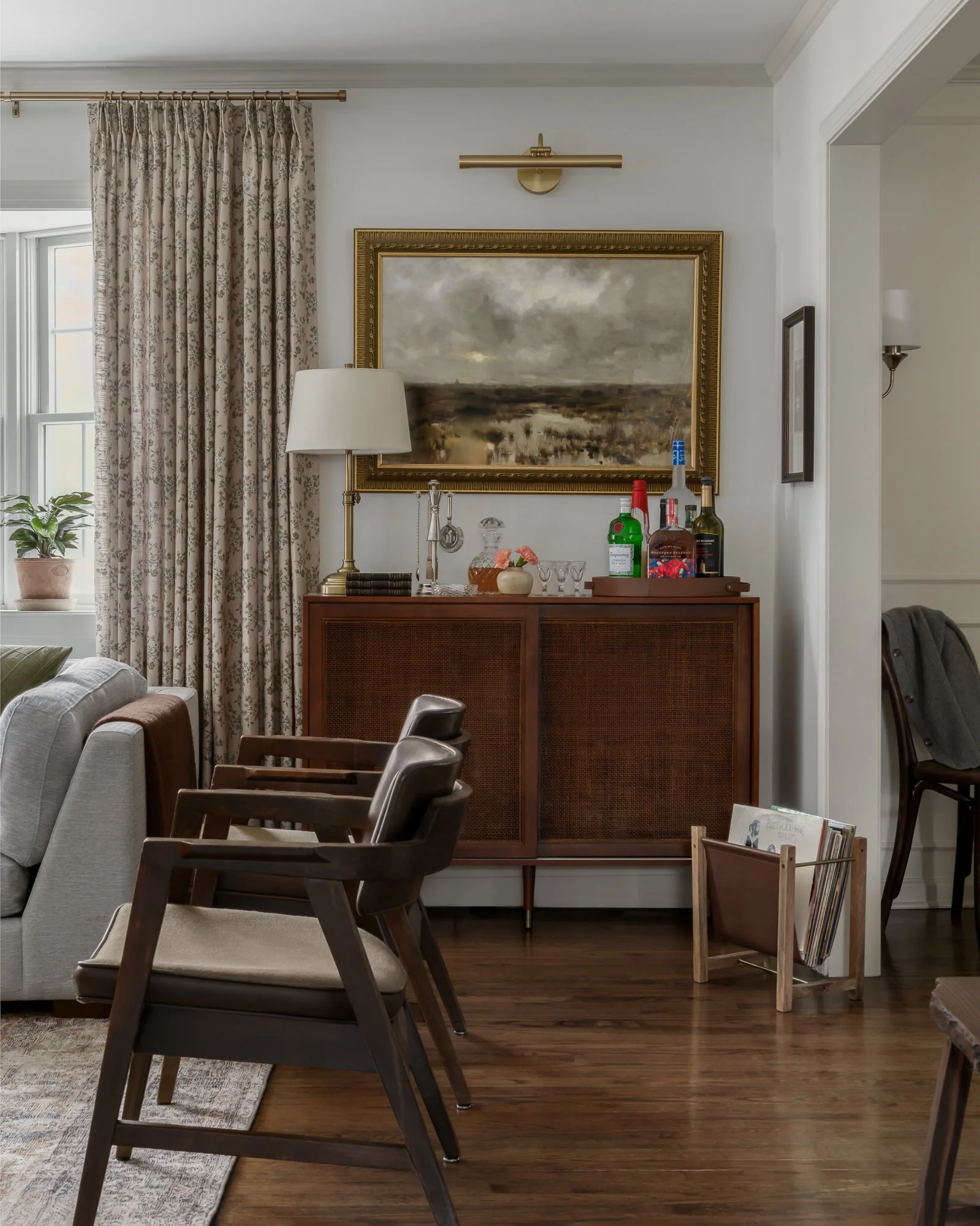 Interior of a living room with a wooden sideboard, a lamp, framed painting, and various bottles on top. There are two dark wooden chairs and a gray sofa, with a window and curtain on the left side, and a magazine rack on the floor.