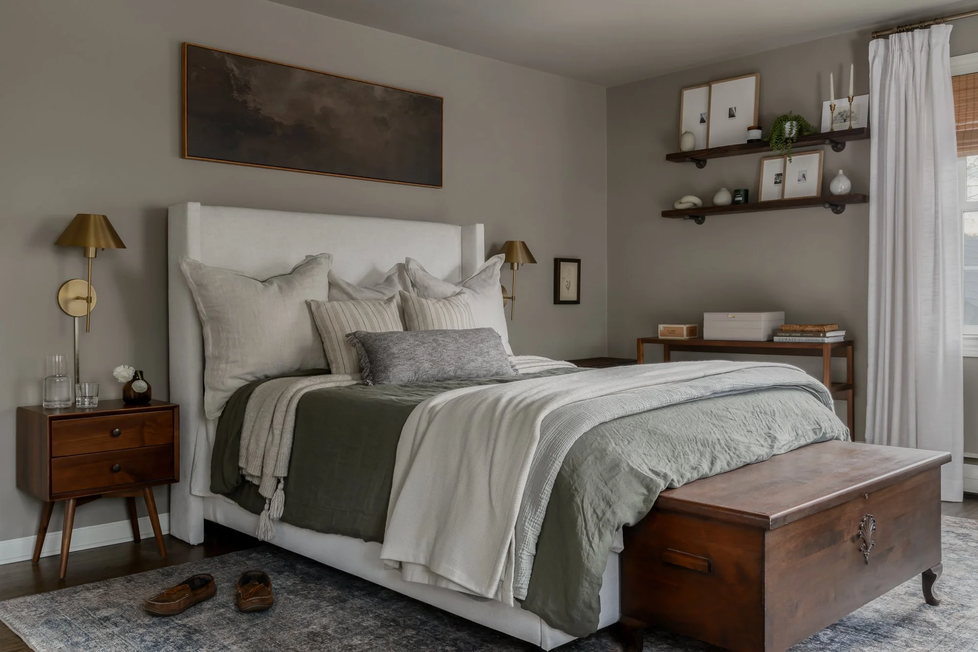 A neatly made bed with multiple pillows, a white headboard, and a green and beige blanket in a cozy bedroom. There are two brass wall-mounted lamps on either side of the bed, a wooden nightstand with glasses and a clock on the left, a framed picture 