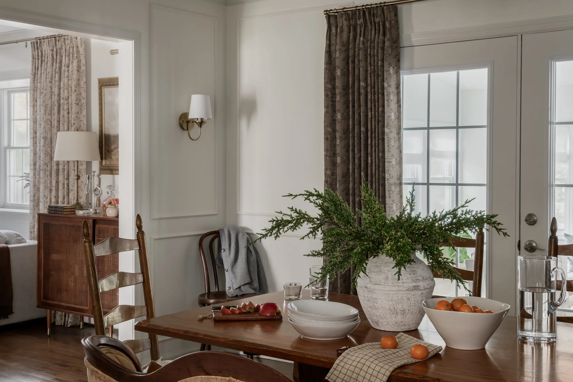 A dining room with a wooden table decorated with a large vase of greenery, bowls of fruit, glass of water, and a pitcher. The room has white walls, dark curtains, and multiple windows allowing natural light.