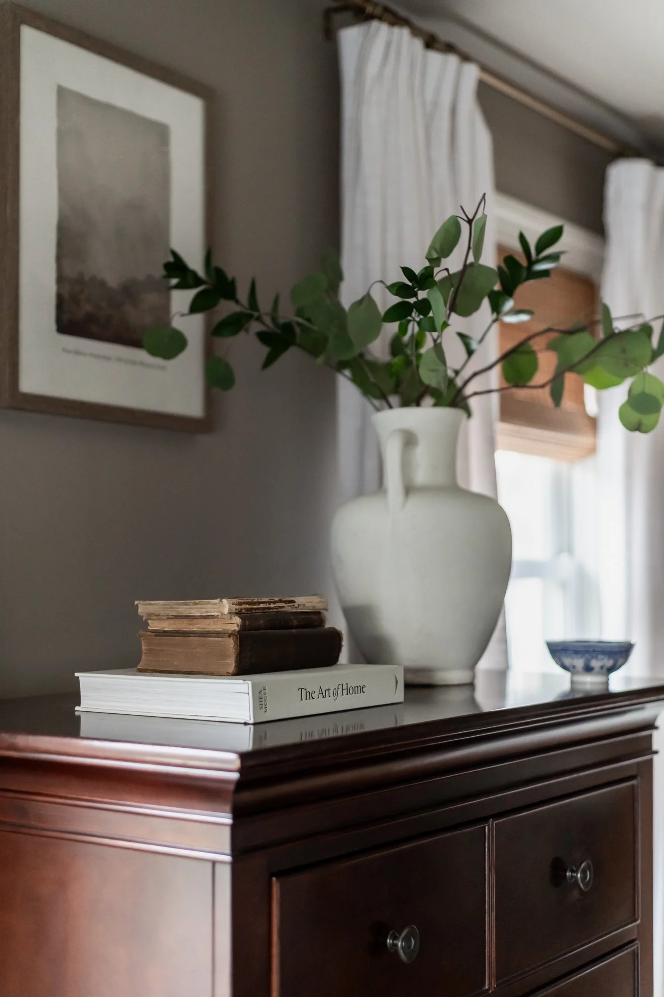 A wooden dresser with three drawers, decorated with a large white ceramic vase containing green leafy branches, three old hardcover books, a white art book titled 'The Art of Home,' and a small blue patterned bowl. There is framed artwork on the wall