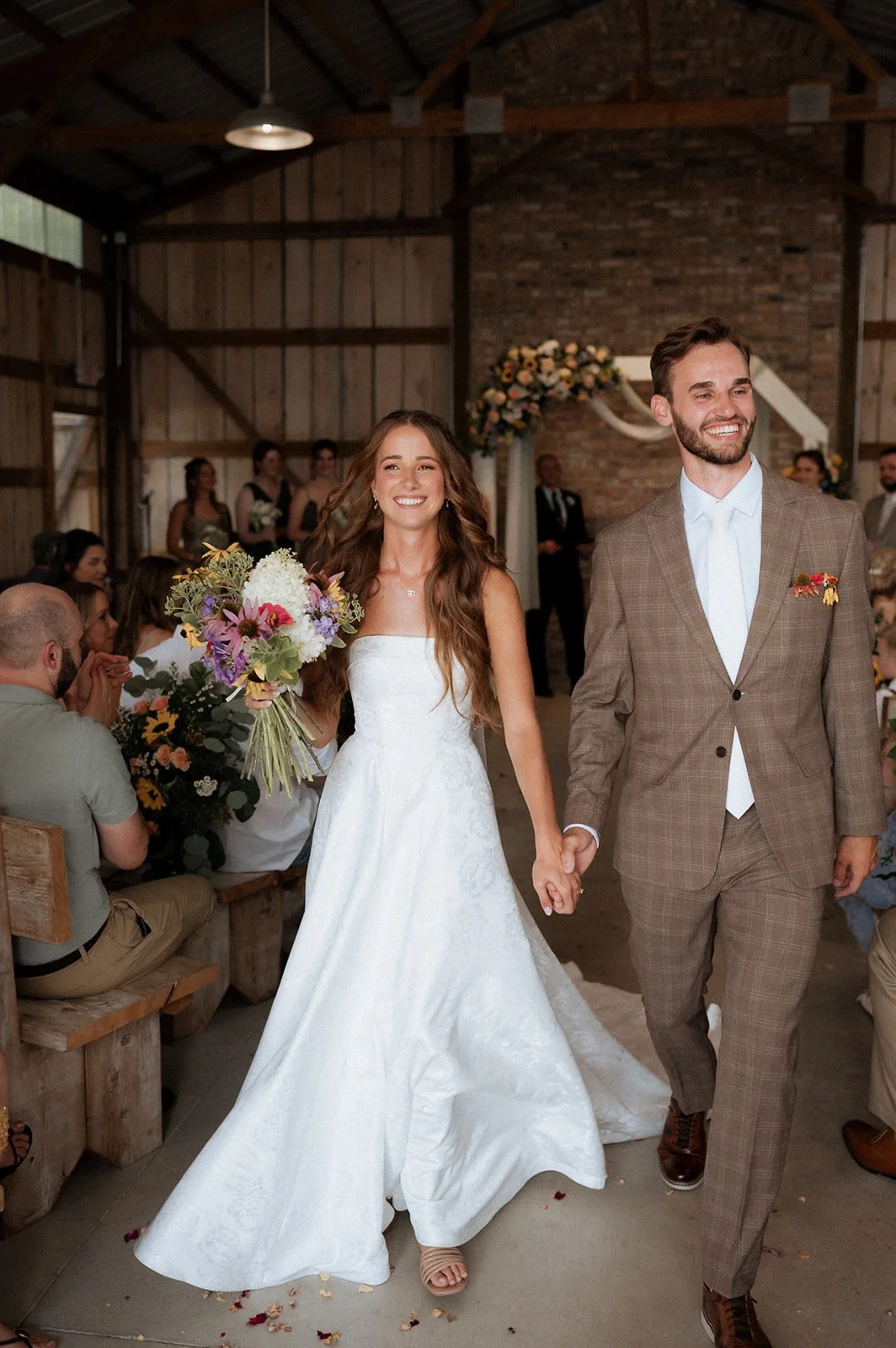 A newlywed couple holding hands and walking down the aisle during a wedding ceremony, with guests seated on either side and a floral arch in the background.
