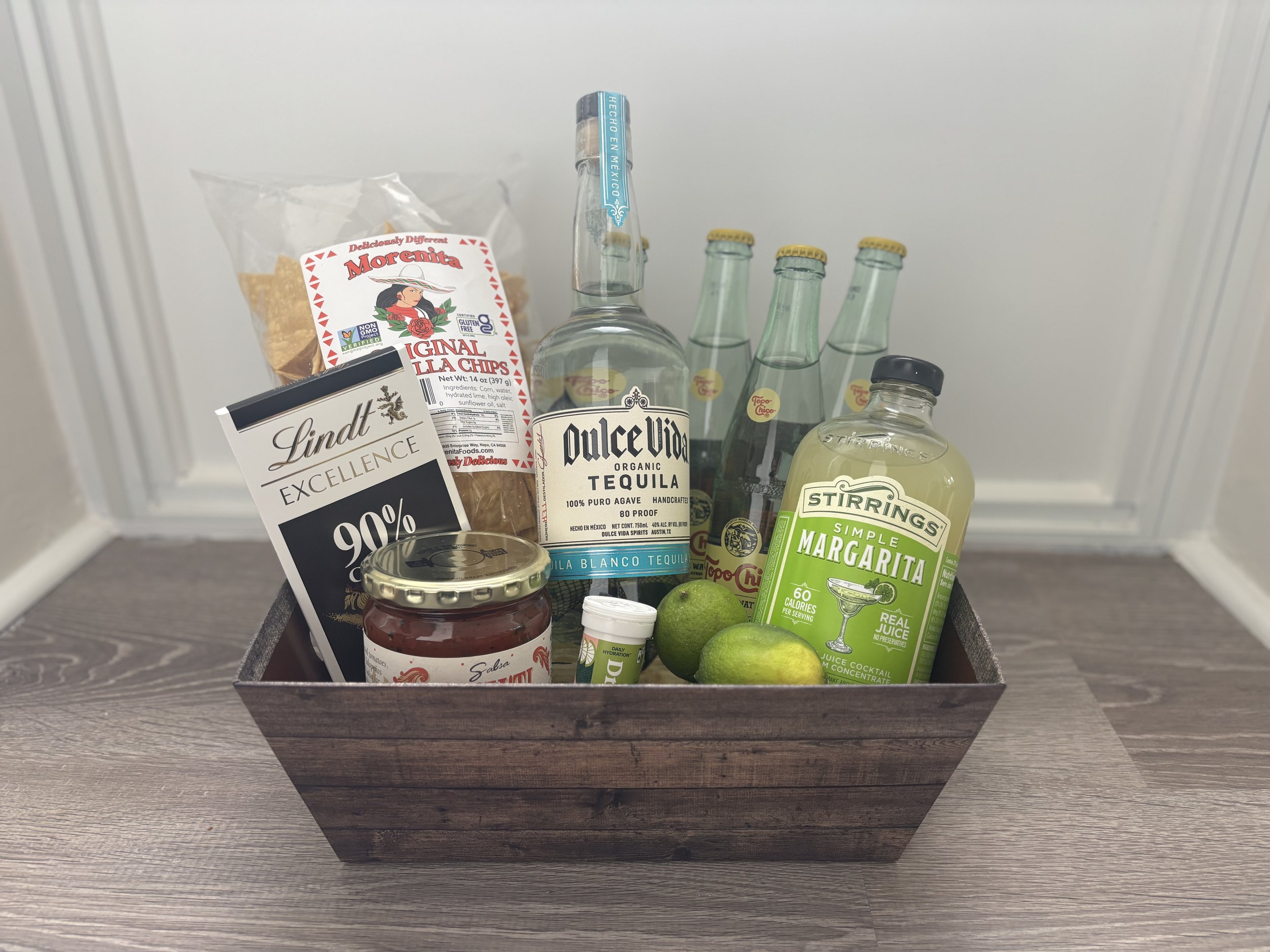 Gift basket with tortilla chips, tequila, margarita mix, club soda, Lindt chocolate, lime, salsa, and shot glass on wooden floor.
