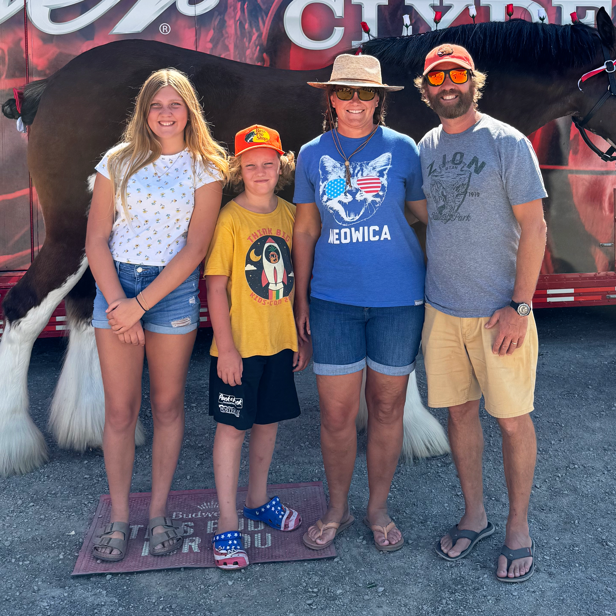 A family of four standing in front of a large horse trailer with a horse visible inside. The family includes a teenage girl in a white floral top and denim shorts, a young boy in a yellow shirt and black shorts, a woman in a blue t-shirt and denim shorts, and a man in a gray t-shirt and beige shorts. They are outdoors on a gravel surface, smiling at the camera.