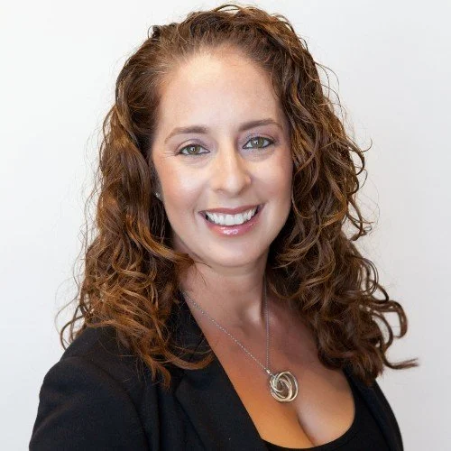 A woman with curly red hair smiling at the camera, wearing a black blazer and a circular pendant necklace against a plain white background.