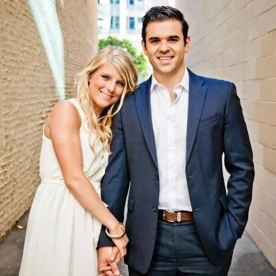 A smiling couple, a woman with long blonde hair wearing a white dress and a man in a dark suit, standing close together outdoors.