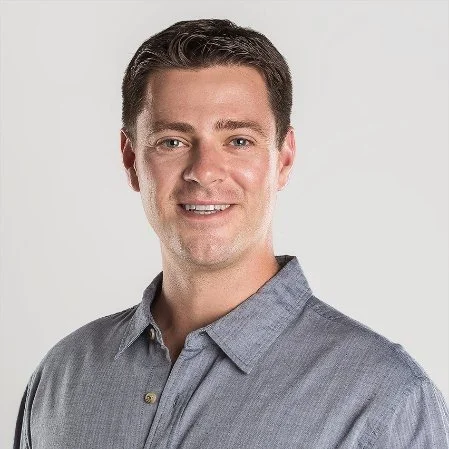 Photo of a man with short brown hair, light skin, wearing a gray collared shirt, smiling against a plain white background.