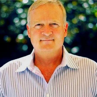 Portrait of a middle-aged man with short blonde hair, wearing a striped collared shirt, outdoors with blurred greenery in the background.