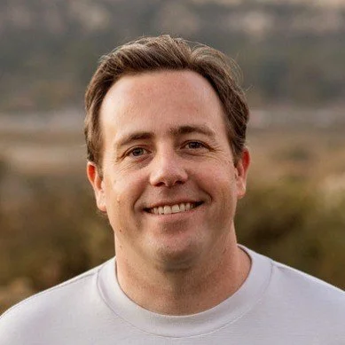 A man with brown hair smiling outdoors with a blurred natural background.