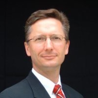 A man with glasses, wearing a suit and tie, smiling against a dark background.