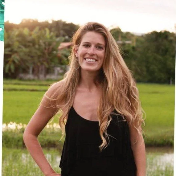 A woman with long wavy blonde hair smiling outdoors in front of a green field and trees during sunset.