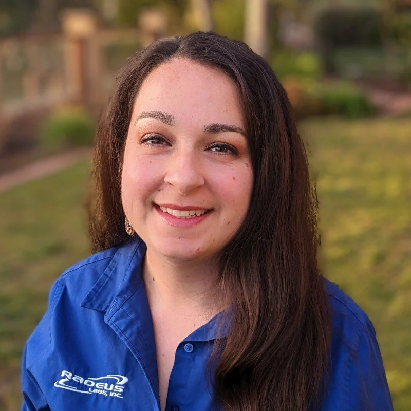 A young woman with long dark hair smiling outdoors in a blue shirt, with greenery and trees in the background.
