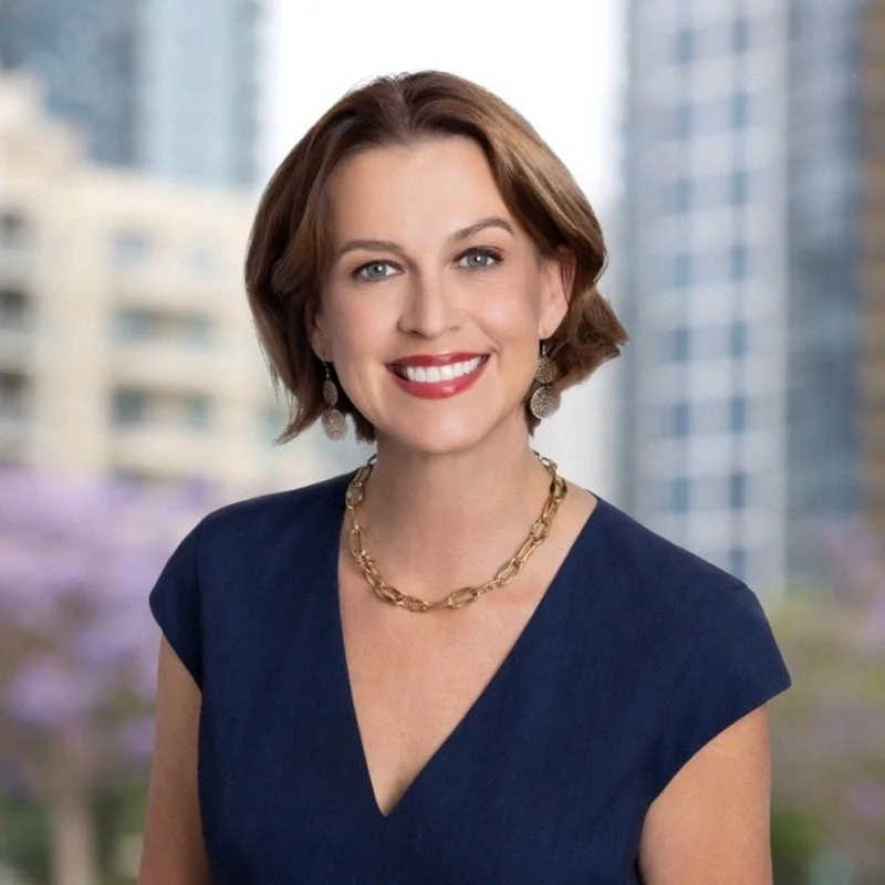 A woman with short brown hair, wearing a navy blue dress and gold jewelry, smiling in front of a cityscape background.