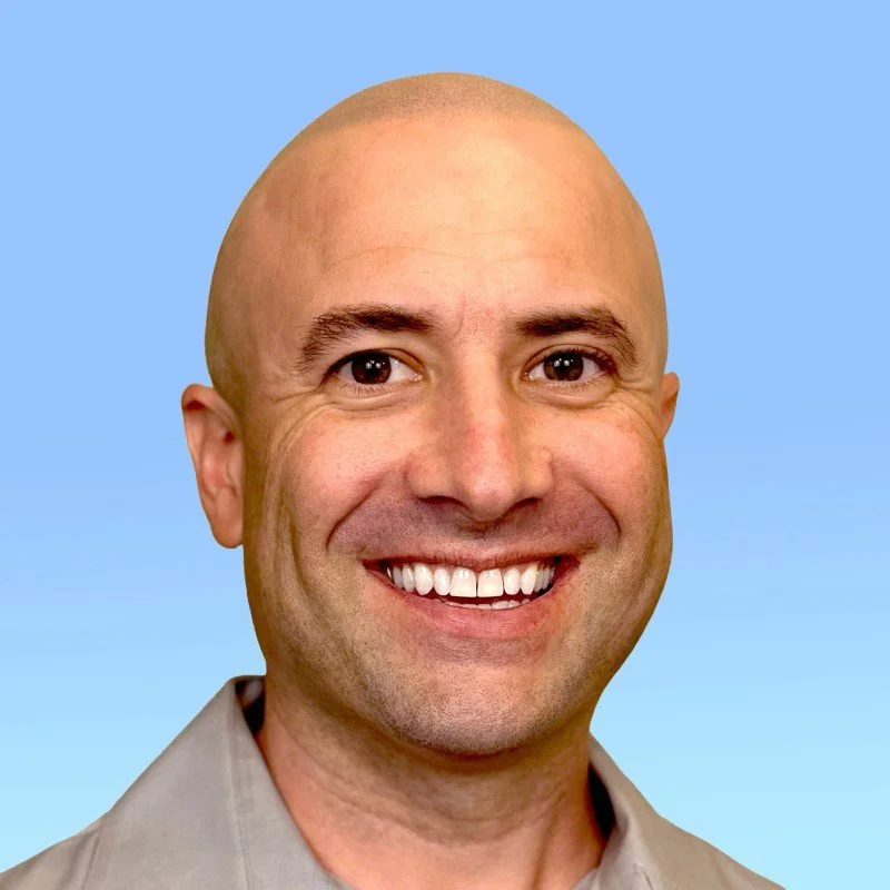 A smiling man with a bald head, brown eyes, and wearing a gray shirt against a blue background.