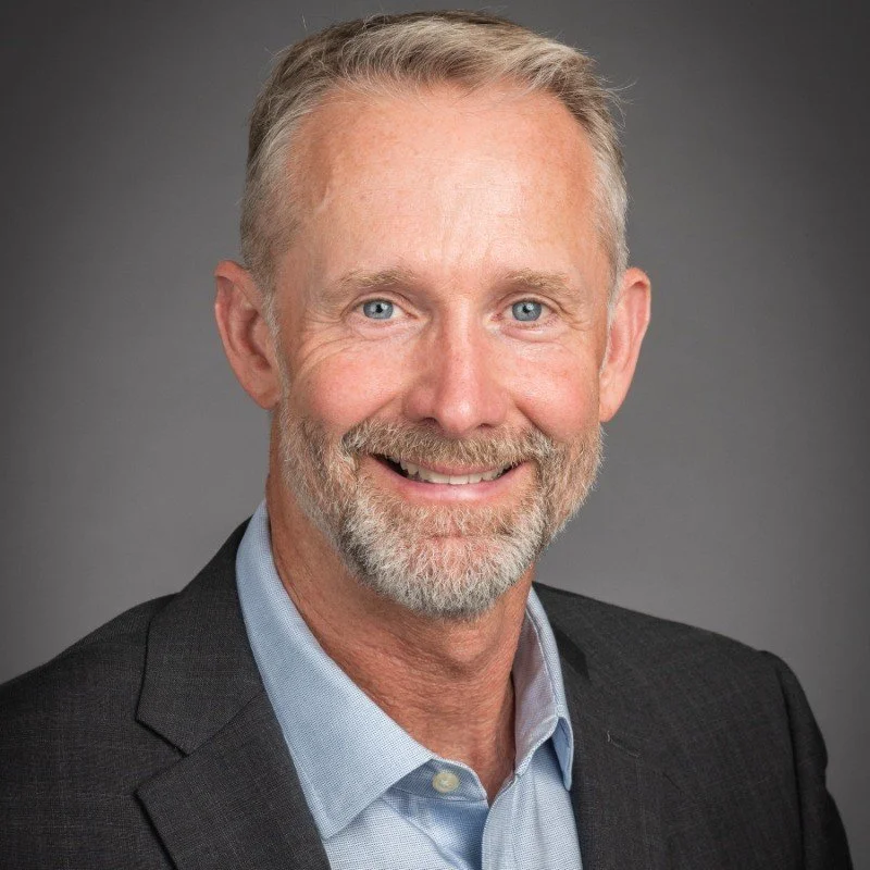 A middle-aged man with gray hair and a beard, smiling, wearing a dark suit jacket and a light blue collared shirt, against a plain gray background.