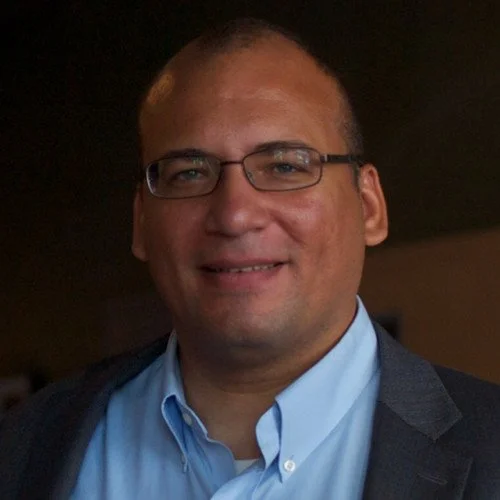 A smiling man wearing glasses, a light blue shirt, and a dark blazer.