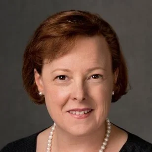 A woman with short, reddish-brown hair, wearing a black top and a pearl necklace, smiling against a gray background.