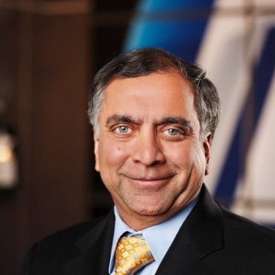 A middle-aged man with gray hair, wearing a black suit, blue shirt, and yellow tie, smiling at the camera.
