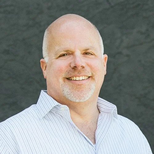 A smiling man with a beard and shaved head, wearing a white striped shirt, standing outdoors in front of a dark gray wall.