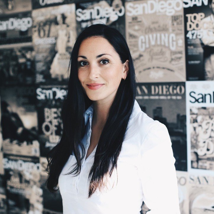 A woman with long black hair and a white blouse standing in front of a collage of San Diego-themed posters and magazines.