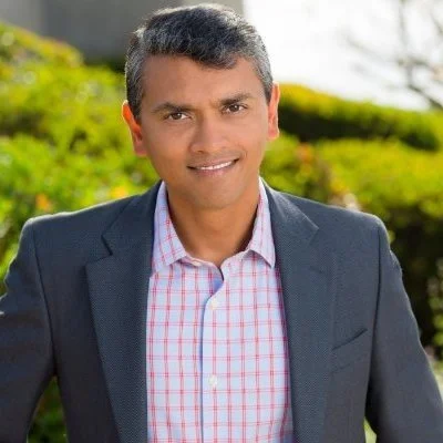 A man smiling outdoors wearing a gray suit jacket and a pink checkered shirt, with greenery in the background.