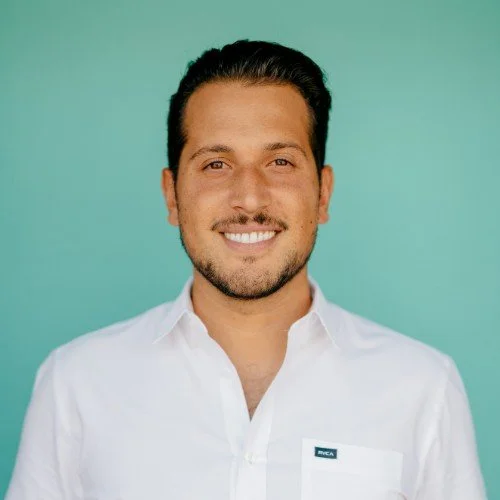 Portrait of a smiling man with dark hair, beard, wearing a white shirt against a teal background