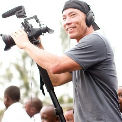 A man smiling, wearing a black cap and headphones, holding a professional video camera on a tripod outdoors.