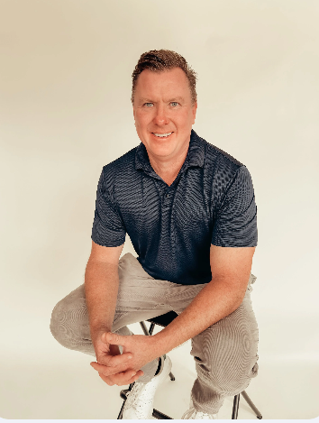 A man with light hair smiling, wearing a light blue polo shirt with arms crossed, against a black background.
