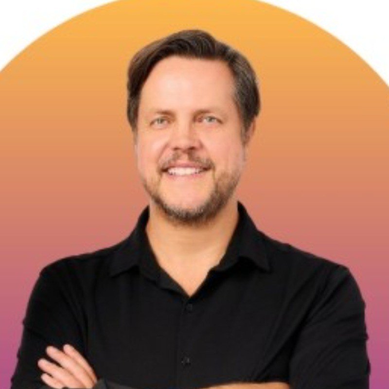A man with brown hair and a beard, wearing a black shirt, standing with arms crossed against a peach and pink gradient background.