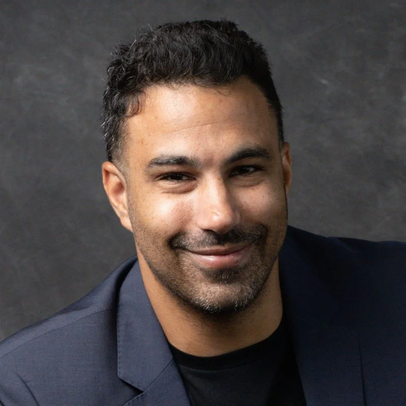A smiling man with dark hair and a goatee, wearing a dark blazer, against a dark textured background.