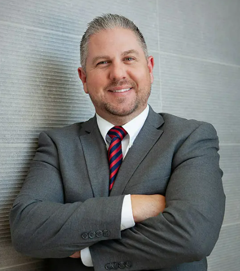 A confident man in a gray suit with a white shirt and a striped red and blue tie, smiling with arms crossed, standing against a textured wall.
