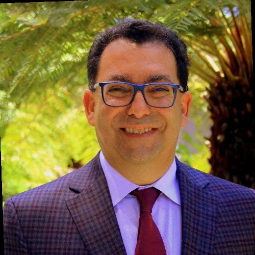 A man wearing glasses, a suit, and a tie, smiling outdoors with green foliage in the background.