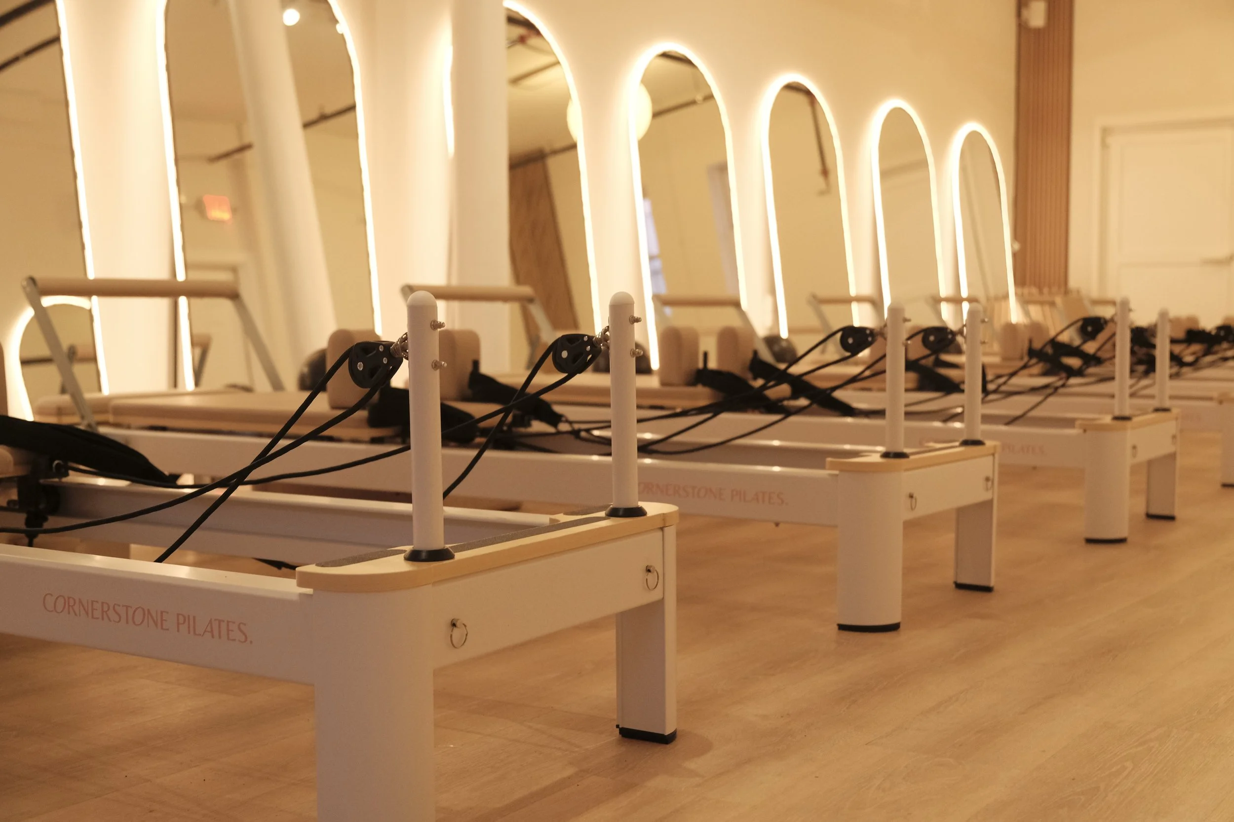 A room with multiple pilates reformer machines arranged in rows, illuminated by soft lighting. The machines have wooden frames, black straps, and pulleys, with the words "Cornerstone Pilates" visible on their sides. The room has wooden floors and walls with a warm tone.
