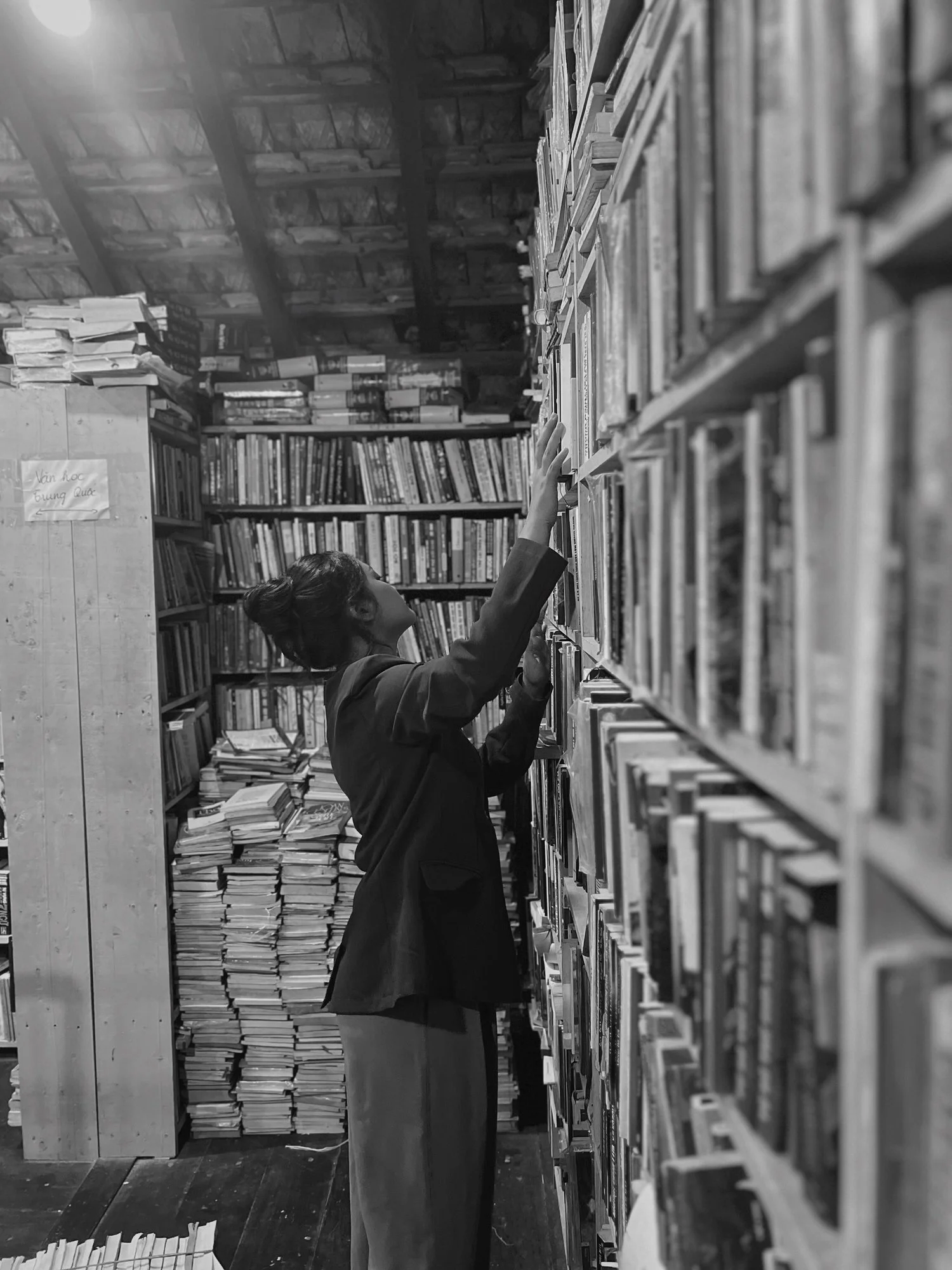 A woman in a bookstore or library reaches for a book on a high shelf surrounded by shelves filled with books and stacked papers